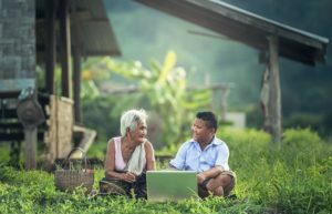 A grandmother and grandson smile lovingly at each other. They sit in front of a computer in a field of greenery.