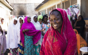 A young Nigerian girls stands smiling the foreground. In the background her friends stand, also smiling