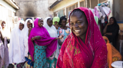 एक जवान नाइजेरियन केटीहरू अग्रभूमिमा मुस्कुराउँदै उभिरहेकी छन्. पृष्ठभूमिमा उनका साथीहरू उभिएका छन्, पनि मुस्कुराउदै