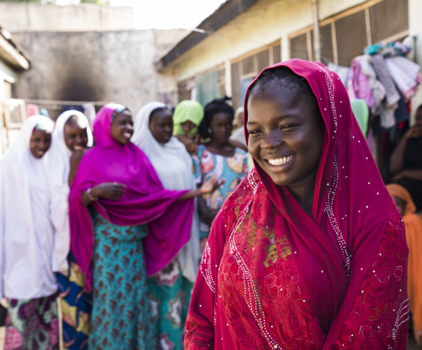 A young Nigerian girls stands smiling the foreground. In the background her friends stand, also smiling