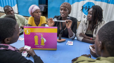 Seorang wanita muda duduk dikelilingi oleh orang muda yang lain. Dia menunjukkan penggunaan kondom dalaman/wanita.
