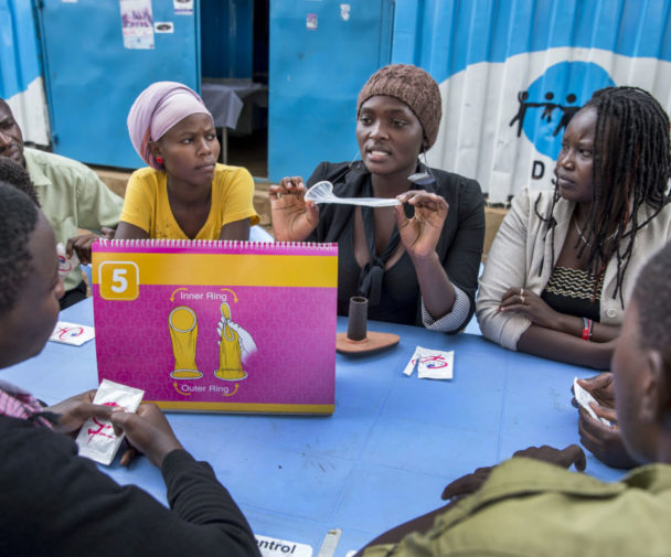 A young woman sits surrounded by other young people. She demonstrates the use of an internal/female condom.