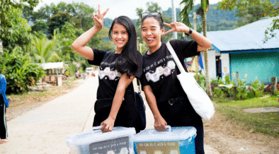 Jovens trabalhadores da saúde em uma estrada em Palawan, Filipinas. Ambos estão vestidos de preto, estão sorrindo para a câmera, e estão segurando suas mãos em um sinal de paz. Ambos também estão carregando caixas de plástico na frente deles.