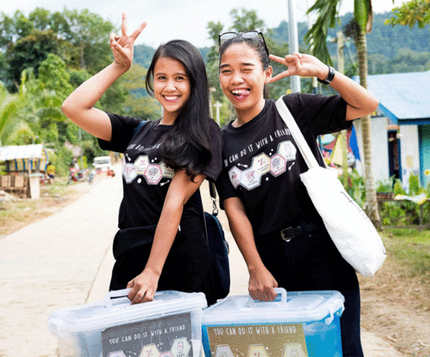 Young health workers standing in a road in Palawan, Philippines. Both are dressed in all black, are smiling at the camera, and are holding up their hands in a peace sign. Both are also carrying plastic boxes in front of them.