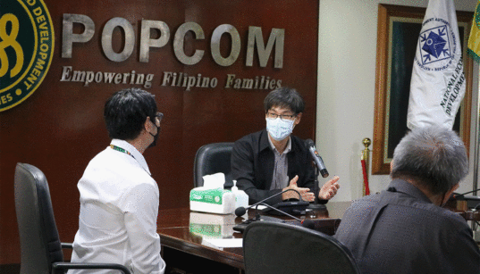 Les employés de POPCOM portant des masques sont assis autour d'une table de conférence pour discuter de leur mandat lors d'une réunion interne. Crédit image: POPCOM
