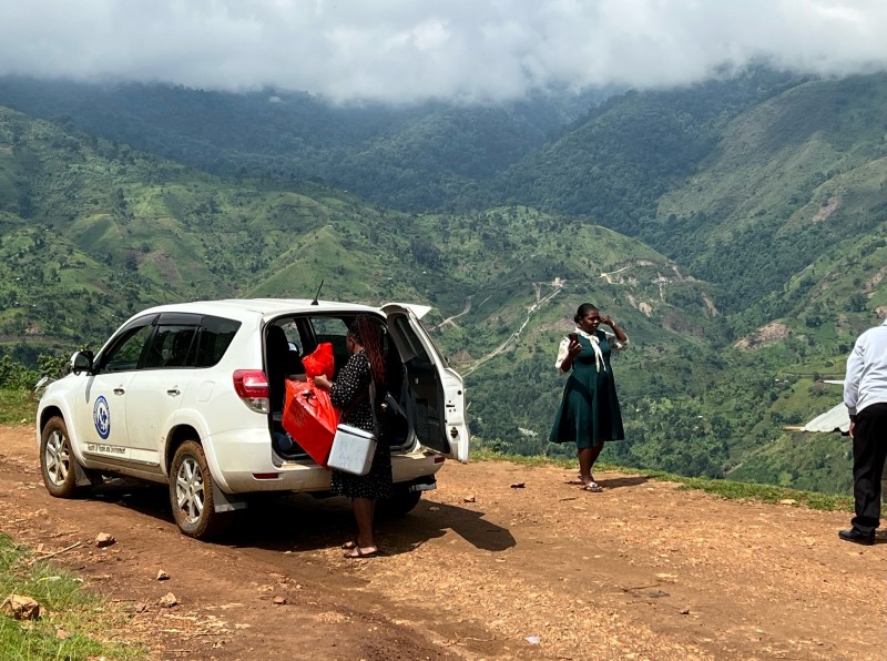 A white vehicle belonging to RCRA Uganda along with several RCRA workers stopping on a dirt road traversing the mountains in Uganda. Photo credit: Rwenzori Center for Research and Advocacy (RCRA)