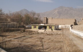 A dirt road to the right of the photo leads to a small green health clinic in the distance. In front of the clinic is a porch area with a group of people sitting outside. Credit: Afghanistan, Department for International Development, 2009.