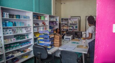 The photo depicts The Kasha Fulfillment Center in Kigali, Rwanda. Kasha is an e-commerce company that enables confidential purchases of health care products, including contraceptives, pregnancy tests, and HIV oral self-test kits. The Kasha Fulfillment Center sells and distributes general beauty and over-the-counter health products. It also relays orders to pharmacies (e.g., for birth control and morning-after pills) for fulfilment and delivery in discreet packaging. The wall to the left in the photo is made of brick and is painted teal. It is lined with white bookshelves with various health and wellbeing products such as soup, shampoo, deodorant, and menstrual pads. There are two white tables in front of these shelves with a horizontal file folder, two computers and a printer on top. There are also two black chairs pushed in, in front of the white tables. A woman sits in the far back left corner of the photo at one of the computers. Another woman stands to the right of the tables and wears a grey t-shirt, jeans, and pink sandals. The wall in the right side of the photo is painted bright pink.