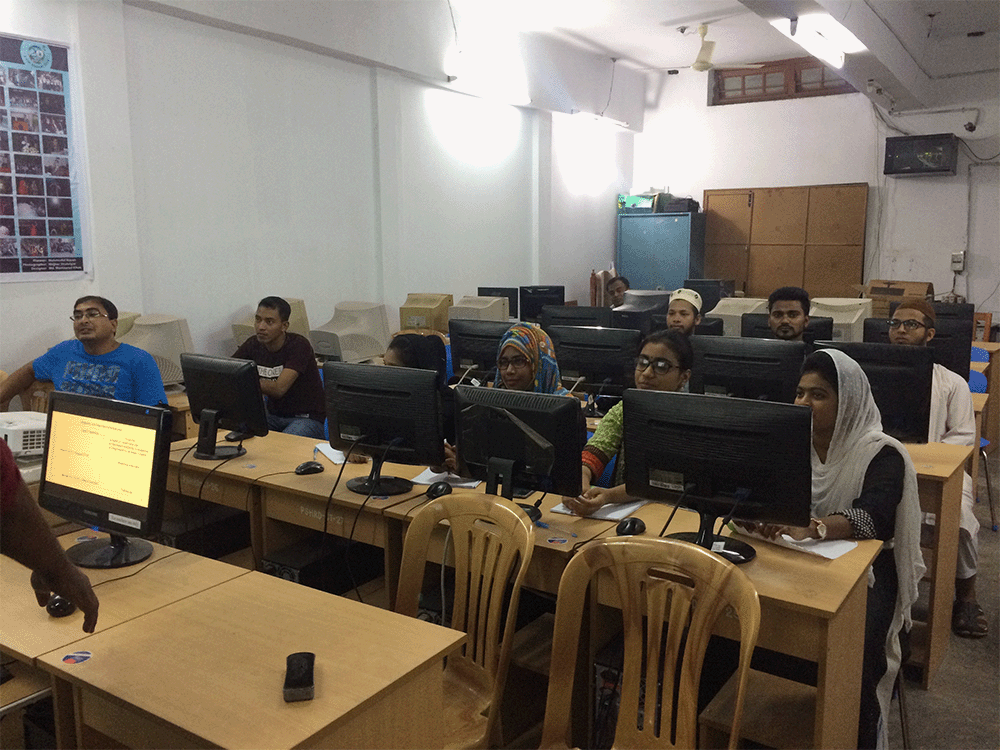 Workshop for Service Provision Assessment survey data handling with the research assistants and the Master course students. A group of ten men and women sit behind rows of wooden desks in front of computers. They sit in a room with white walls and a tall teal cabinet in the back. A person stands at the front of the room behind a wooden table and computer, instructing the research assistants and Master course students. Photo Credit: Bangladesh Research Team, Bangladesh.