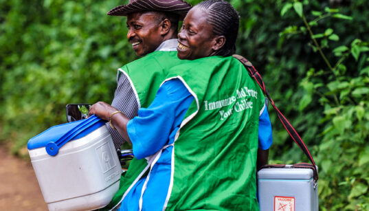 two health workers, part of a resilient supply chain, smiling on a motorcycle delivering vaccines in coolers