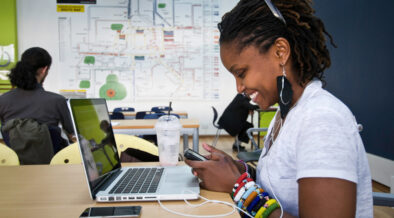 A young Kenyan woman is sitting at a desk with a laptop computer and is holding a cell phone while wearing earphones