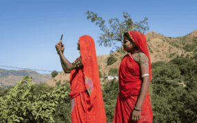 Two women, one holding a mobile phone, beside a small mountain in India.