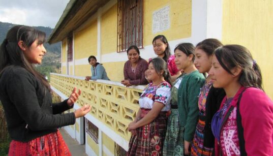 A group of adolescent girls stand outside a yellow building in Guatemala.